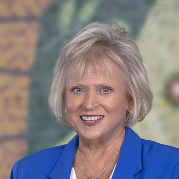 A smiling older woman, Debbie Compton, with short, light blonde hair wears a bright blue blazer and white top, posed in front of a softly blurred, multicolored background.
