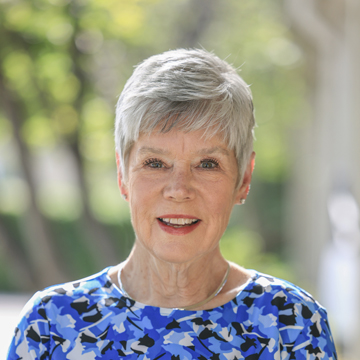 An older woman with short gray hair smiles warmly, wearing a blue patterned top and a silver necklace. She stands outdoors with green trees and soft sunlight in the blurred background.
