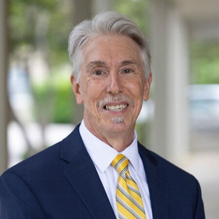 Fred DeMagistris, a smiling older man with gray hair and a goatee, stands outdoors in a navy suit, white shirt, and yellow striped tie, framed by a blurred backdrop of trees and buildings.