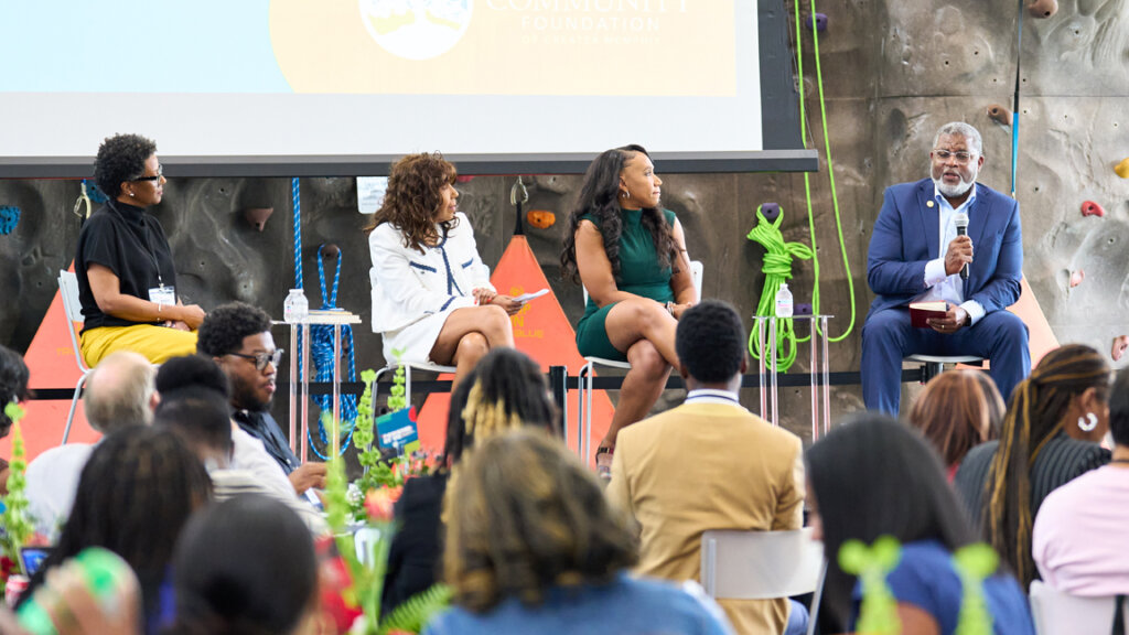 Four panelists sit on stage in front of an audience, engaged in a discussion about building community at an indoor event with a rock climbing wall in the background. One man speaks into a microphone while the others listen.