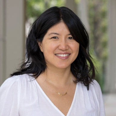 A woman with medium-length black hair, wearing a white blouse and a gold necklace, smiles outdoors with a blurred background of columns and greenery.