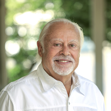 An older man with white hair and a beard smiles while wearing a white collared shirt, standing outdoors with a blurred green and white background.
