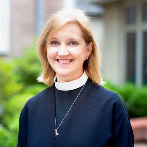 A smiling woman with shoulder-length blonde hair, identified as Katherine Bush, wears a black clerical robe and white collar as she stands outdoors in front of a blurred building and greenery.