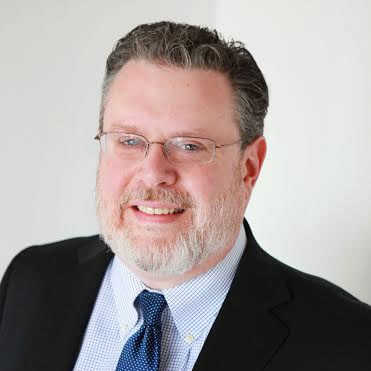A man with short brown hair, glasses, and a gray beard is smiling. He is wearing a dark blazer, light blue checkered shirt, and a blue tie against a plain light background.