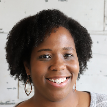 Ruth Abigail Gardner, a woman with natural curly hair, smiles at the camera. She is wearing dangling earrings and a light top, with a white background behind her.