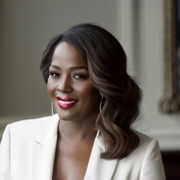 Schanaque Watson, with wavy dark hair and red lipstick, smiles at the camera in a softly lit, elegant room while wearing a crisp white blazer.
