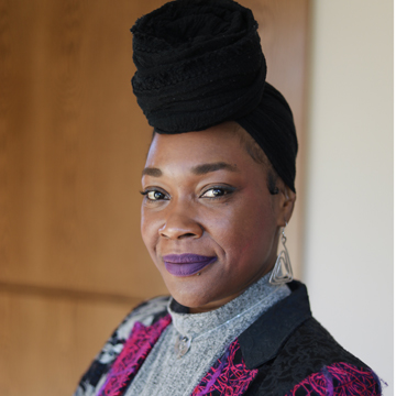 Shanette Parks, with a confident expression, wears a black headwrap, dangly earrings, a gray high-neck top, and a dark jacket with pink patterns, standing indoors against a blurred wooden background.