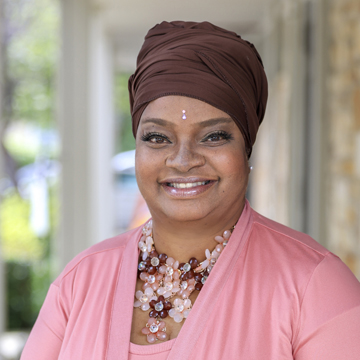 Tyra Turner-Cleasant wears a brown headwrap, pink top, and beaded flower necklace as she smiles at the camera. A small decorative bindi adorns her forehead. The softly blurred background features greenery and a building.