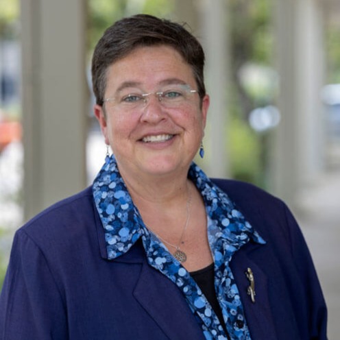 A smiling person with short dark hair and glasses wears a blue patterned shirt under a dark blue blazer, standing outdoors with blurred columns and greenery in the background.