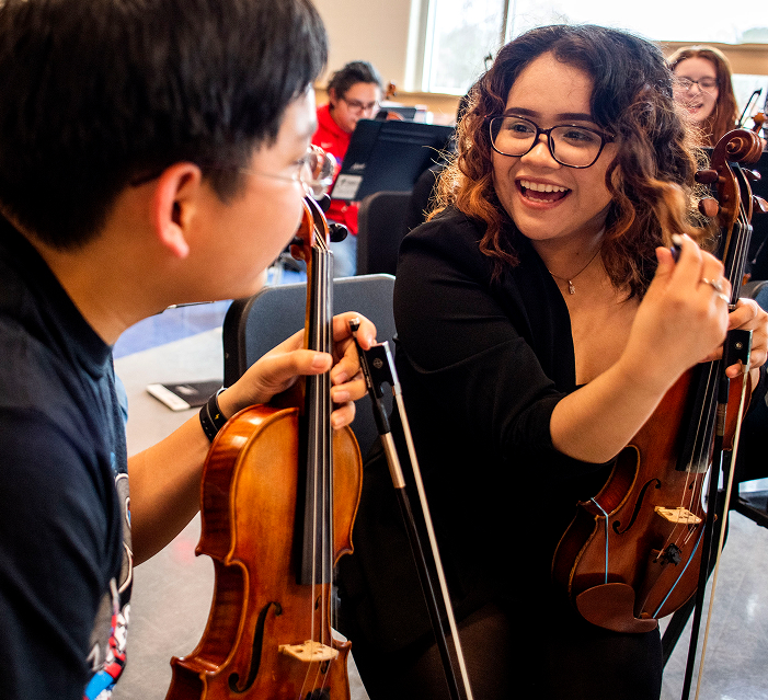 Two young musicians with violins smile and talk to each other during a rehearsal in a music room, with other orchestra members visible in the background.