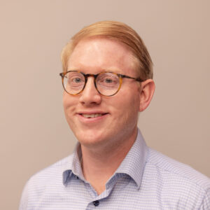 A man with short, light brown hair and glasses is smiling. He is wearing a blue and white checkered shirt, and the background is plain and beige.