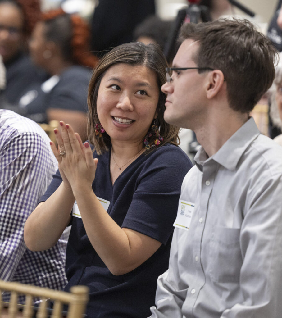 A woman smiling and clapping while talking to a man in glasses at an indoor event. Both wear name tags and are seated among other people.