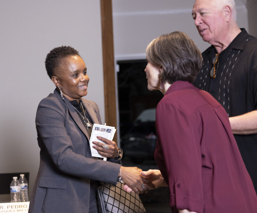 A woman in a suit smiles and shakes hands with another woman in a maroon jacket, while a man in a black shirt stands beside them, all engaged in conversation indoors.