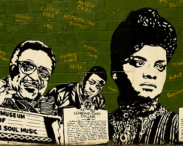 A mural painted on a green brick wall features three prominent Black figures, including a woman with an elaborate hairstyle and two men, one with glasses. Yellow text and names are scattered in the background.