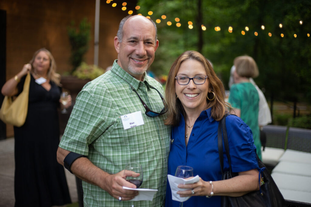 A man in a green plaid shirt and a woman in a blue blouse smile at an outdoor event, each holding a drink. String lights and other people are visible in the background.