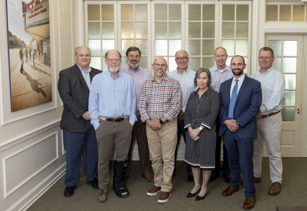 Nine people, eight men and one woman, stand in a row inside a well-lit room with large windows and white walls. Dressed in business casual attire, they smile at the camera, reflecting a team focused on performance and investment.