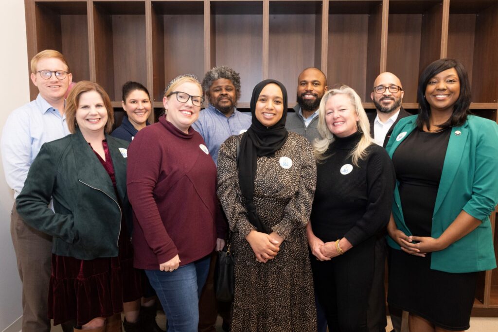 A diverse advisory team of ten adults, smiling together indoors in front of empty wooden shelves, poses for a group photo. Most are wearing business or business-casual attire, showcasing their professional staff unity.