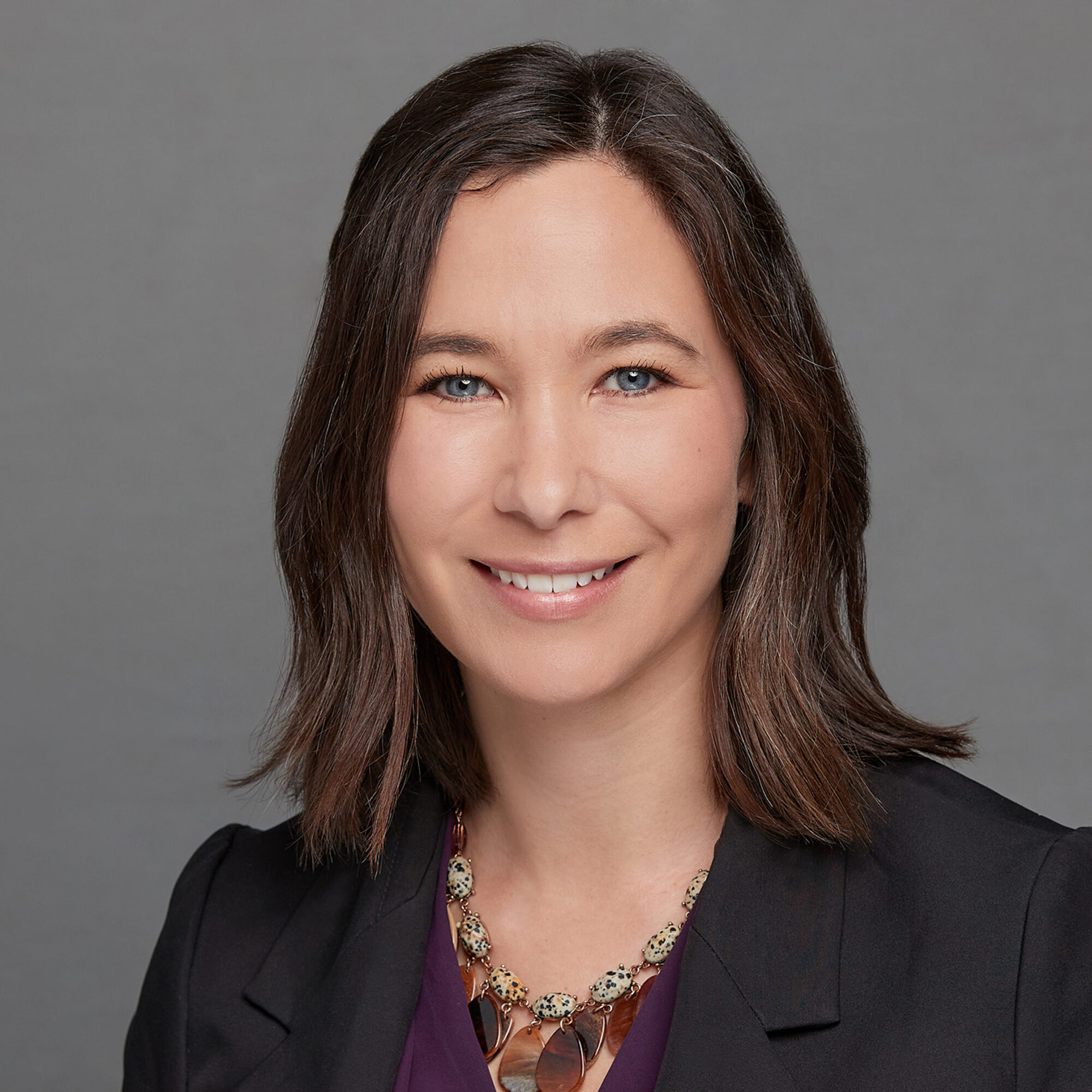 A woman with straight brown hair and blue eyes smiles at the camera. She is wearing a black blazer, a purple top, and a beaded statement necklace, with a plain gray background behind her.