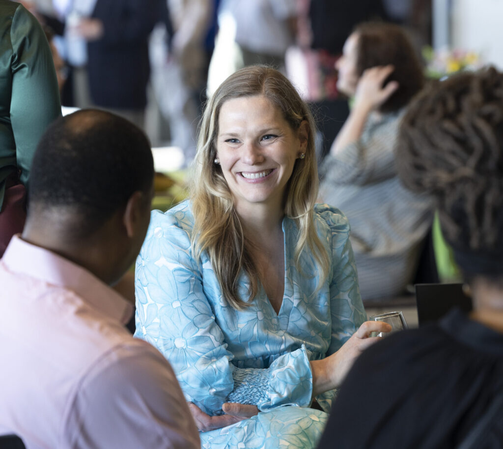 A woman in a light blue dress smiles while chatting with two people at an indoor social event. She holds a drink, appears engaged in conversation, and shows how easy it is to get involved with others. Blurred guests mingle in the background.