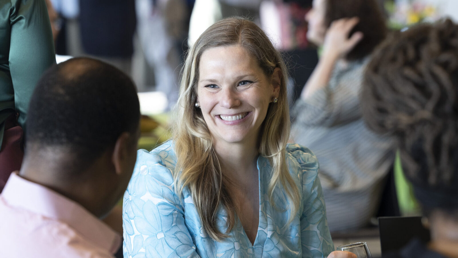 A woman with long blonde hair wearing a light blue patterned blouse smiles while talking to others, encouraging everyone to get involved in the lively, indoor social setting.