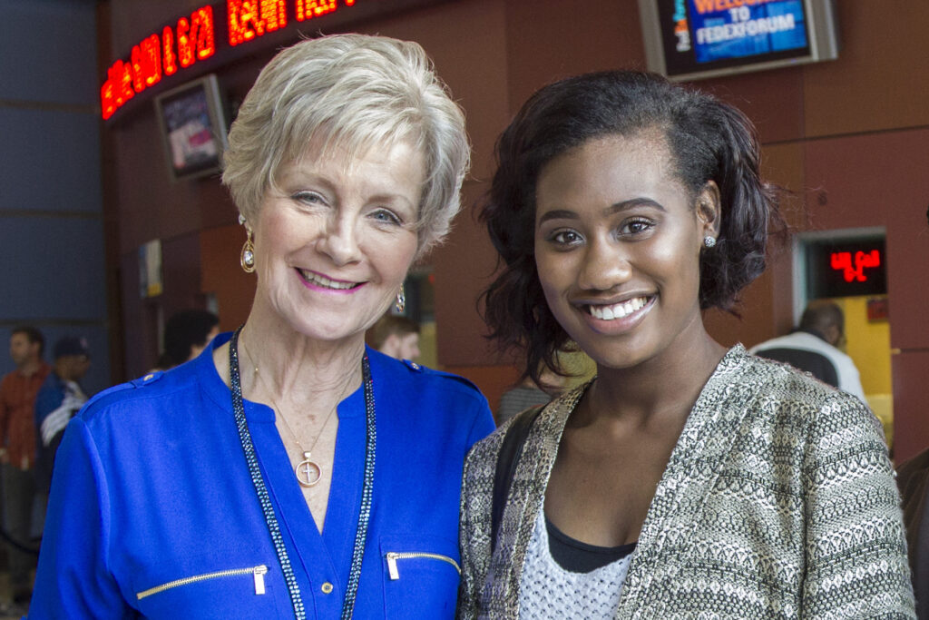 Two women smiling and posing together indoors, with a blurred background featuring digital signs about scholarships and other people. One woman has short blond hair and wears a blue top; the other has shoulder-length dark hair and wears a patterned jacket.