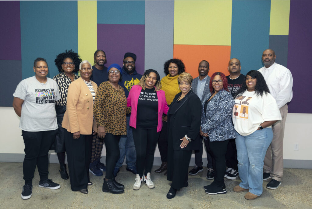 A diverse group of fifteen adults stand together indoors, smiling at the camera. The background features a colorful geometric wall with blocks in blue, green, yellow, orange, and purple—perfect for gathering inspiration or to Submit Feedback.