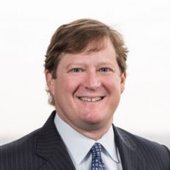 A man with light brown hair wearing a dark pinstripe suit, light blue shirt, and patterned tie, smiling at the camera against a plain, light background.