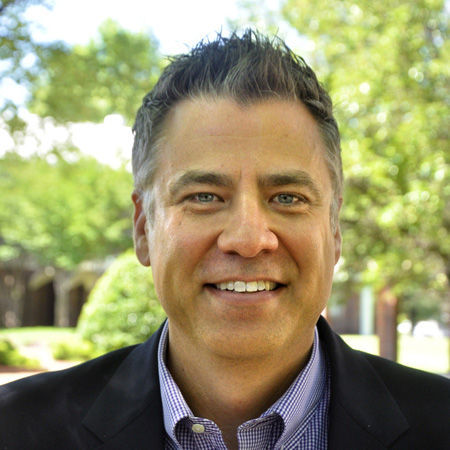 A man with short, dark hair smiles outdoors. He is wearing a dark blazer and a checked shirt. Green trees and shrubs are blurred in the sunny background.
