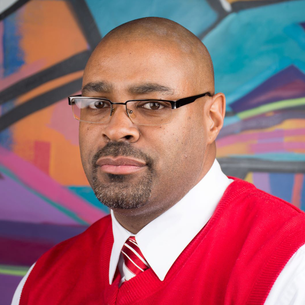 A man wearing glasses, a white shirt, a red vest, and a red striped tie poses in front of a colorful abstract mural.