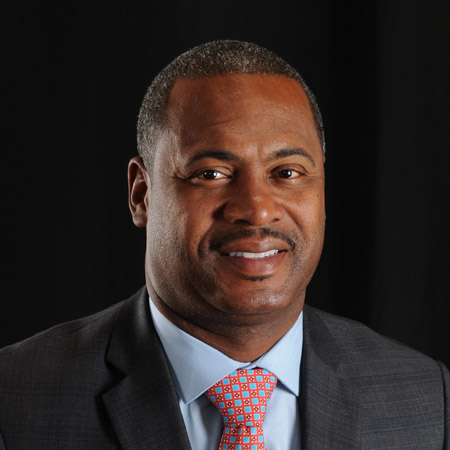 A man wearing a gray suit, light blue shirt, and patterned orange tie smiles slightly in front of a black background.