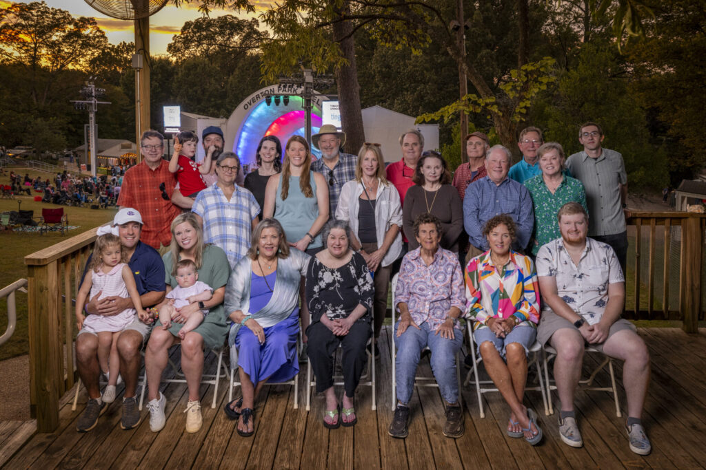 A large group of people of various ages pose for a photo on a wooden deck outdoors, with trees and a colorful event stage in the background. Some are seated in front, while others stand behind them, smiling at the camera.