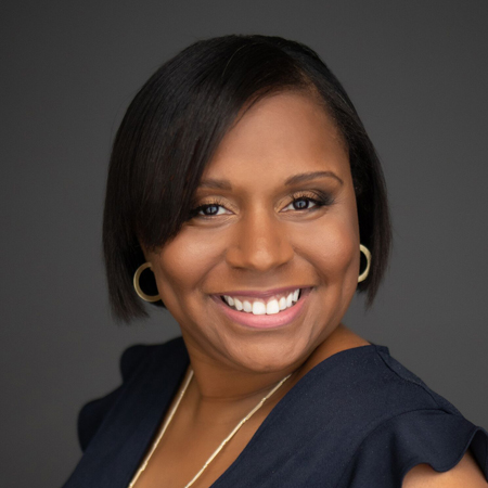A woman with short, straight black hair smiles at the camera. She is wearing gold hoop earrings, a navy blue top, and a gold necklace, with a neutral dark gray background.