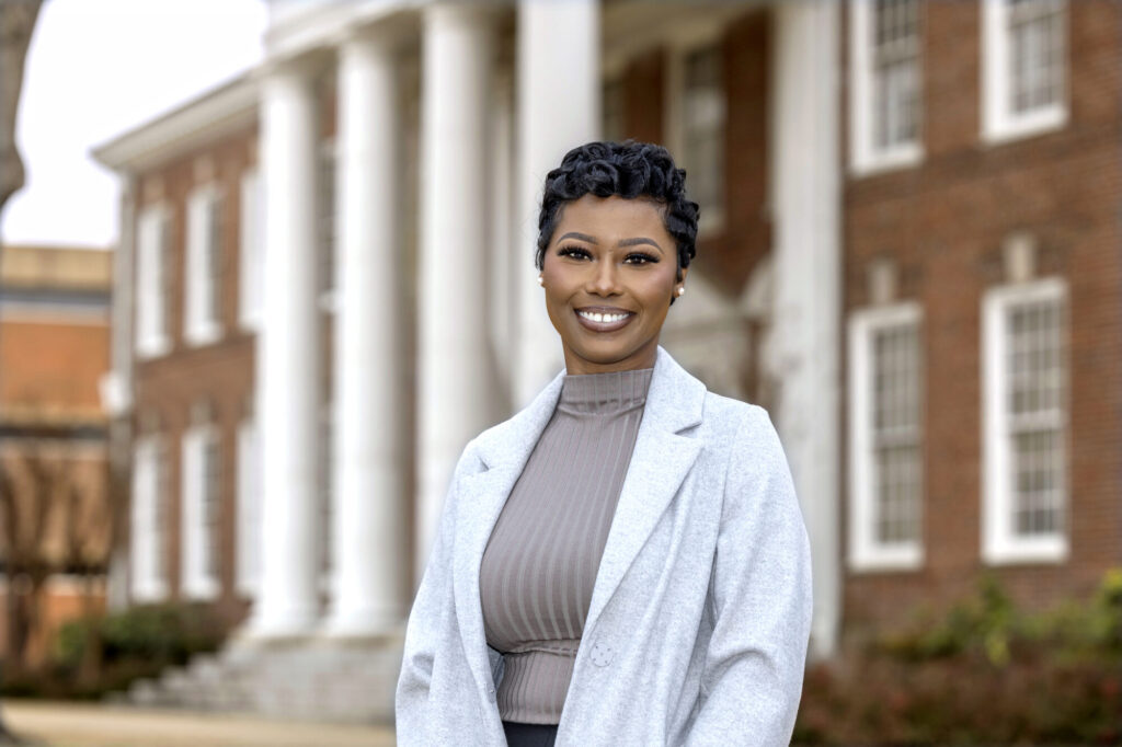 A woman with short black hair, wearing a light gray coat and a ribbed turtleneck, stands smiling in front of a red-brick building with white columns—perhaps celebrating news about scholarships or grants. The background is slightly blurred.