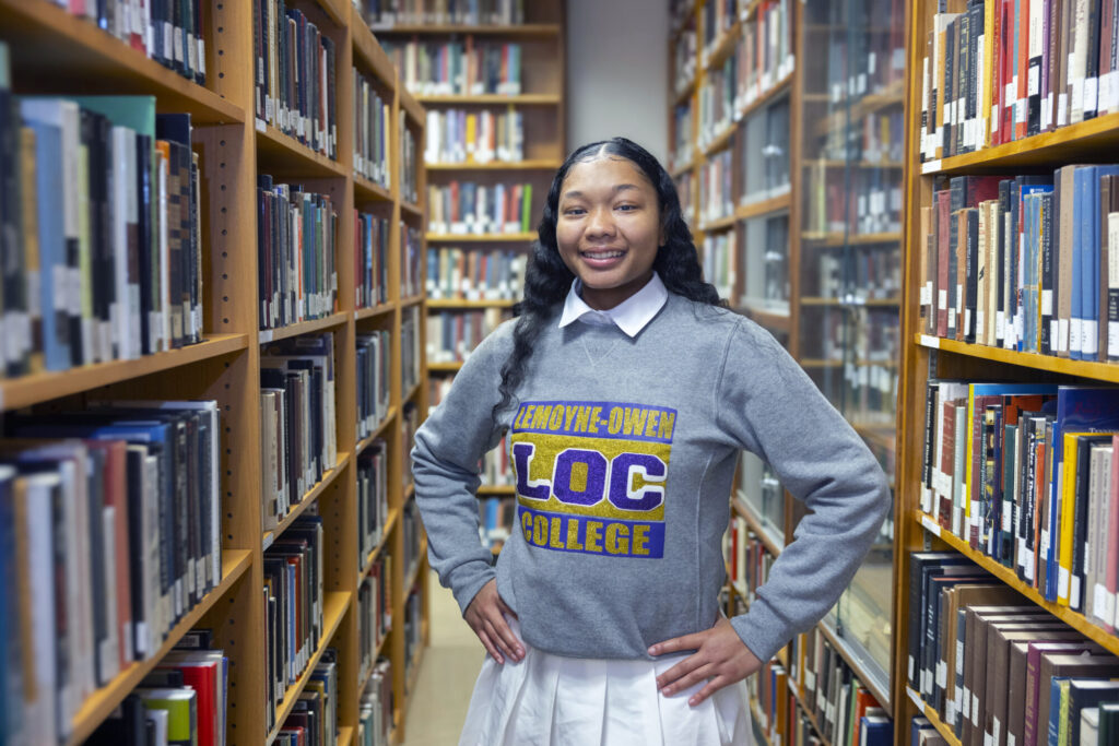 A young woman stands confidently between bookshelves in a library, smiling and wearing a gray Lemoyne-Owen College sweatshirt and white skirt, surrounded by rows of books—a testament to the opportunities offered through scholarships at the college.