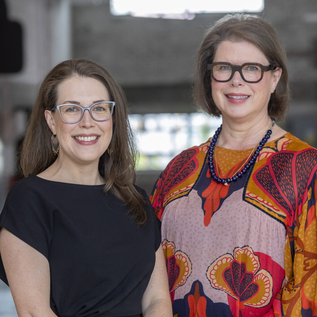 Two women with glasses smile side by side indoors. One wears a black dress, the other a colorful patterned dress with a blue necklace. A bright window and interior space are blurred behind them as they discuss philanthropic services.