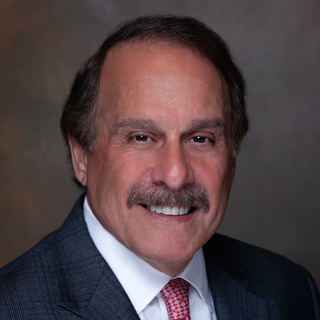 A middle-aged man with dark hair and a mustache, wearing a gray suit, white shirt, and red tie, smiles in front of a neutral, blurred background.