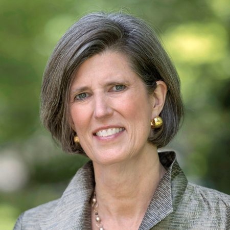 A woman with short gray hair, wearing gold earrings and a collared jacket, smiles outdoors with a blurred green background.