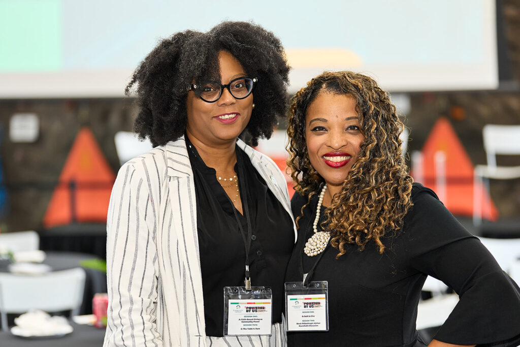 Two women smiling at a professional event, both wearing name badges. One has shoulder-length curly hair and a black top; the other has natural hair and wears glasses and a white striped blazer. Blurred conference setting in the background.