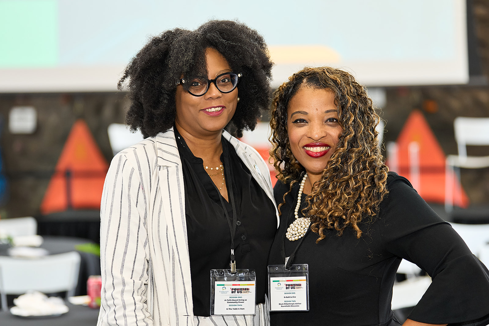 Two women smiling at a professional event, both wearing name badges. One has shoulder-length curly hair and a black top; the other has natural hair and wears glasses and a white striped blazer. Blurred conference setting in the background.