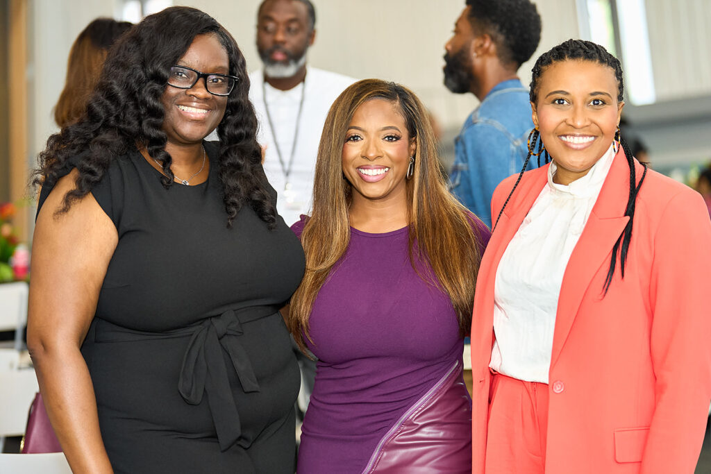 Three women smiling and posing together indoors at an event; two women wear dresses (one black, one purple), and one wears a coral suit over a white blouse. Several people are visible in the blurred background.