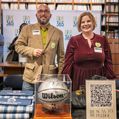 A man and a woman stand smiling behind a table with books, blankets, a basketball, and a QR code sign for Give 365 donations. Bookshelves and a Give 365 banner are in the background.