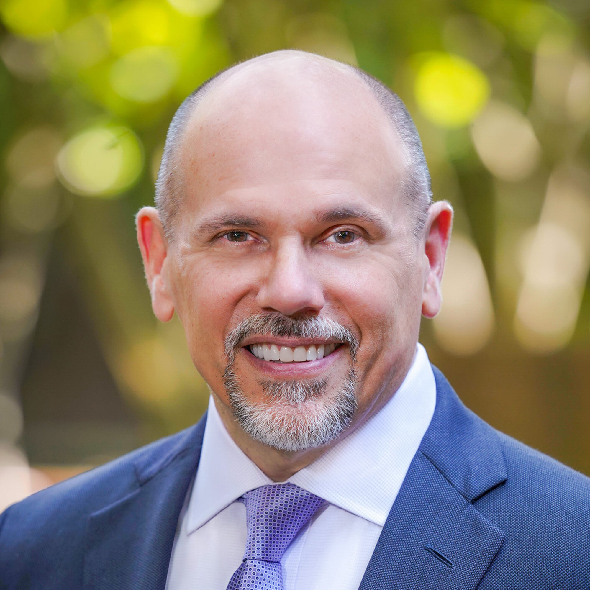 A smiling bald man with a goatee wearing a blue suit jacket, white shirt, and light purple tie stands outdoors with a blurred green background.