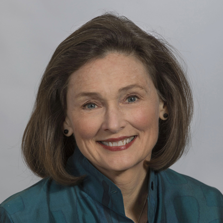 A woman with shoulder-length brown hair and blue eyes smiles at the camera. She is wearing a teal blouse with black earrings and has a light complexion. The background is plain and light gray.