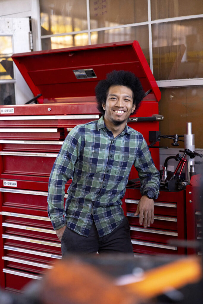 A man with curly hair, wearing a green plaid shirt, smiles while leaning on a red tool chest in a workshop. The background features tools and a window with natural light, creating an inspiring space to learn about scholarships and grants.
