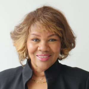 A woman with short, wavy brown hair and blonde highlights smiles at the camera. She is wearing a black blazer, gold hoop earrings, and a gold necklace, with a light-colored background.