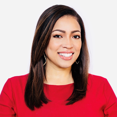 Smiling woman with straight brown hair, wearing a red top and gold earrings, posing against a plain white background.