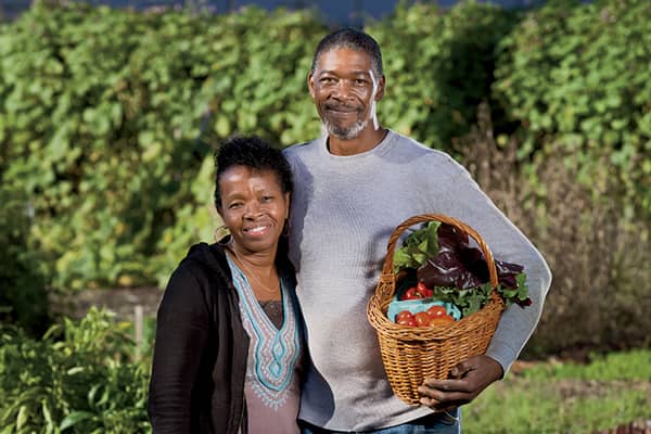 A man and woman holding a basket of vegetables, celebrating the fresh produce grown with the help of local grants.