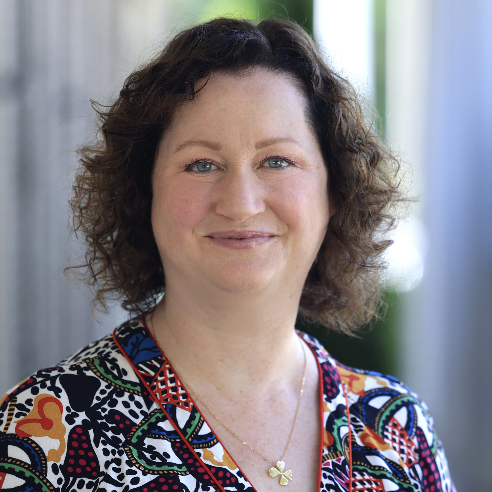 A woman with curly brown hair, wearing a colorful patterned top and a gold necklace with a butterfly pendant, smiles softly while standing in front of a blurred outdoor background.