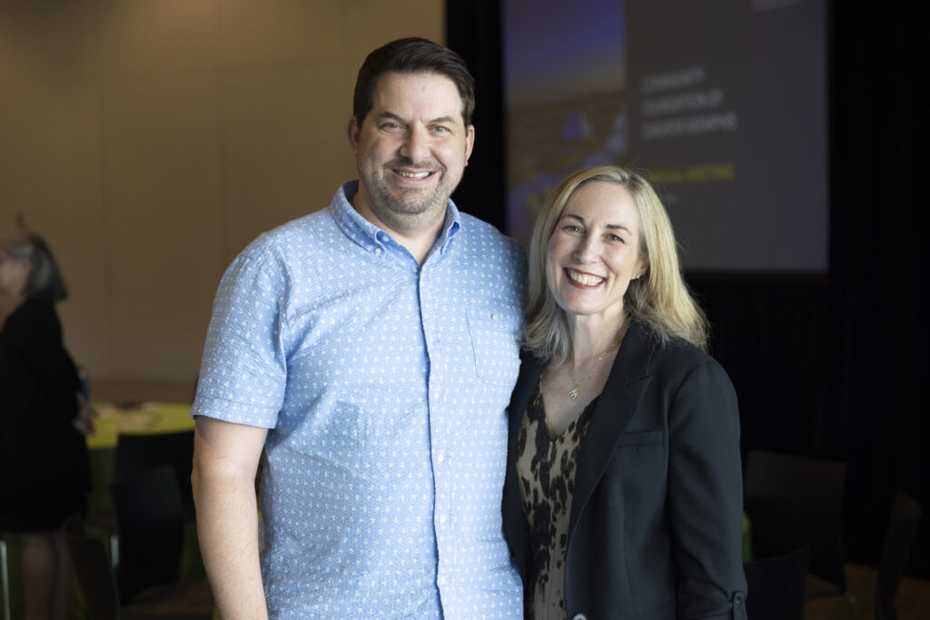 A smiling man in a light blue short-sleeve shirt stands next to a smiling woman in a blazer, posing together indoors with blurred people and a presentation screen in the background.