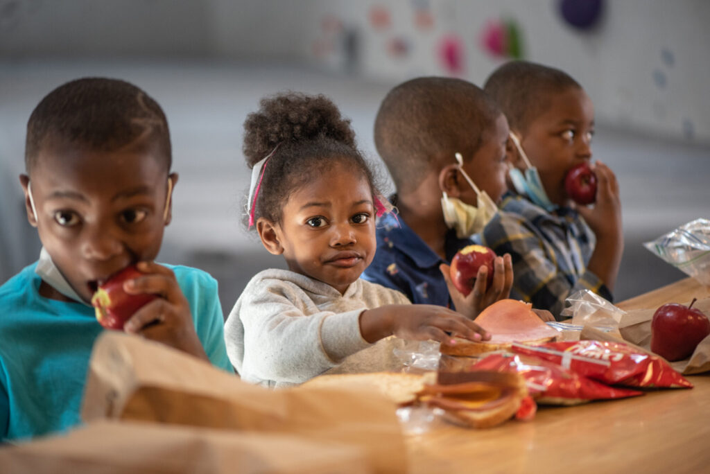 Four young children sit at a table eating apples and snacks. Three of them are looking towards the camera while one looks to the side. Food items and paper bags are spread out on the table in front of them.